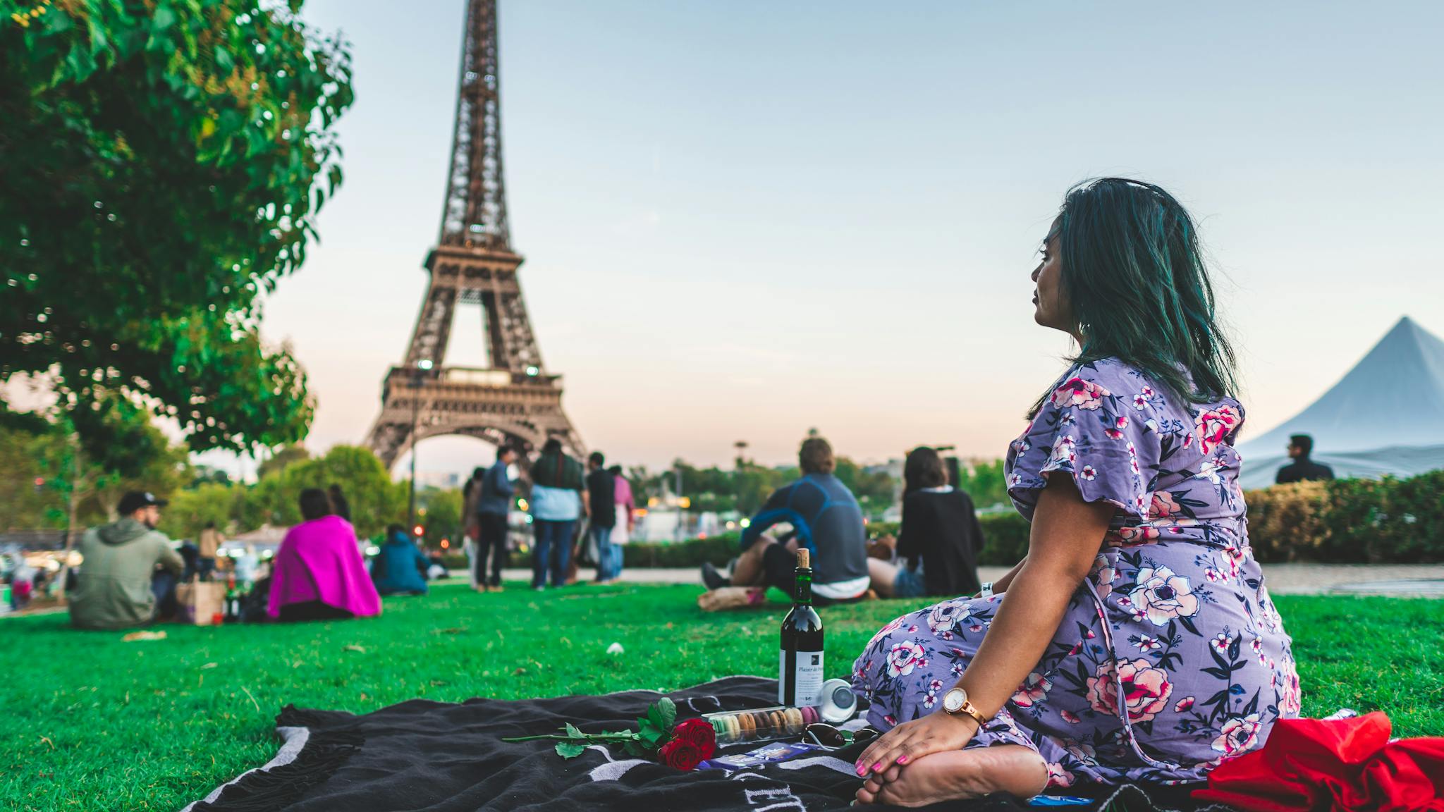 A woman enjoys a leisure picnic with wine in front of the Eiffel Tower on a sunny day in Paris.
