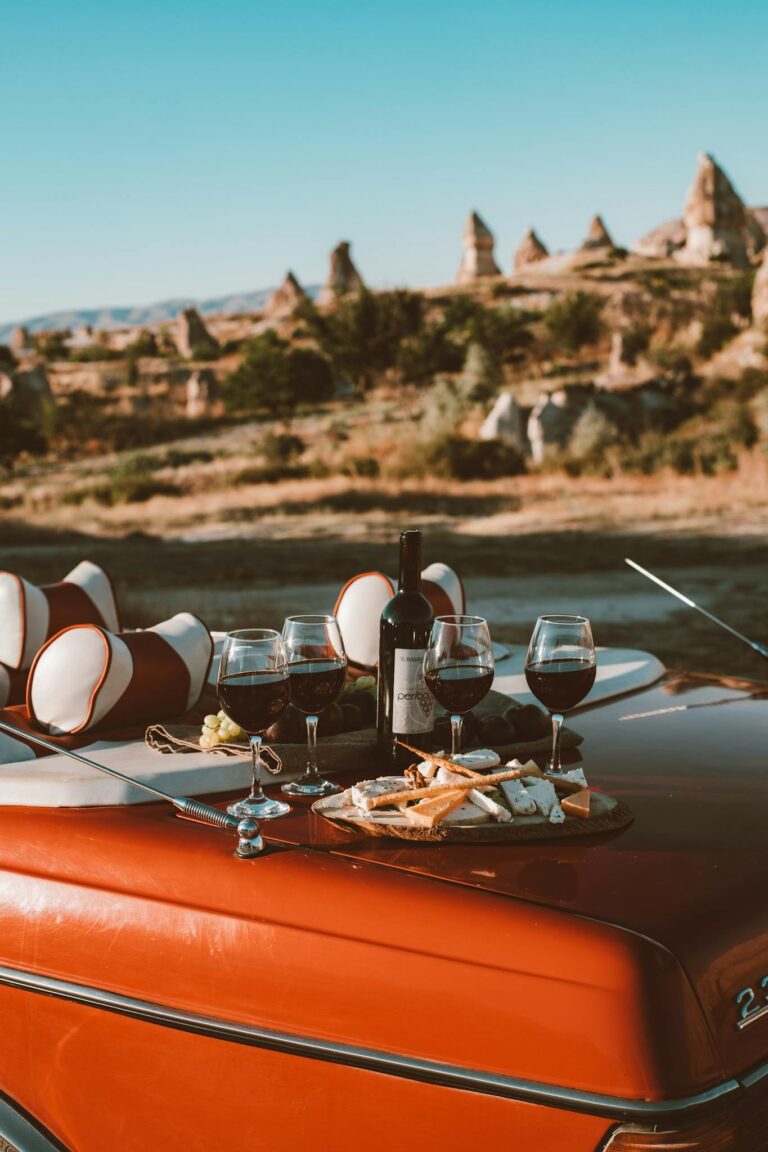 Wine, glasses, and snacks on a classic car outdoors with scenic views.