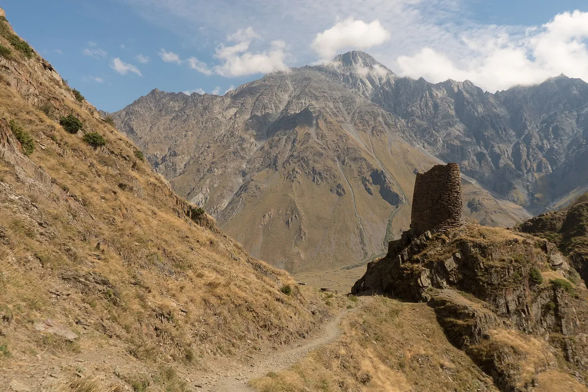 A walking path along a dry mountainside past an old stone tower, with the peaks of Kazbegi National Park behind, Georgia