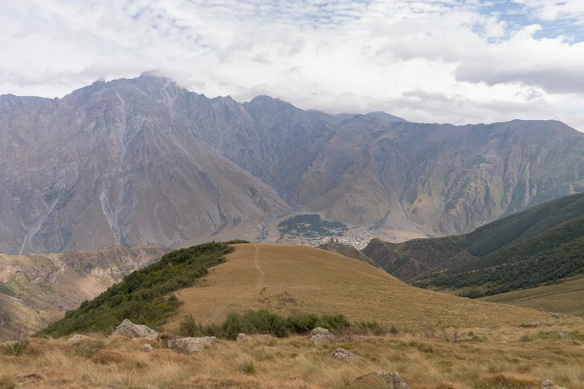 The valley of Stepantsminda seen from a grassy ridge, with the bare peaks of the Kazbegi range behind, Georgia